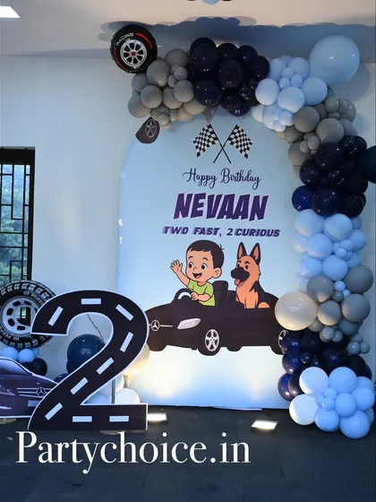 A "Two Fast, 2 Curious" car-themed birthday party. This setup includes a large number '2' cutout designed as a racetrack, a custom backdrop, and a balloon arch in shades of blue, black, and grey.
