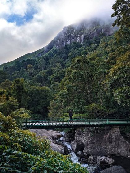 Crossing a bridge over a rushing river in Wayanad, with mist-clad mountains towering above.