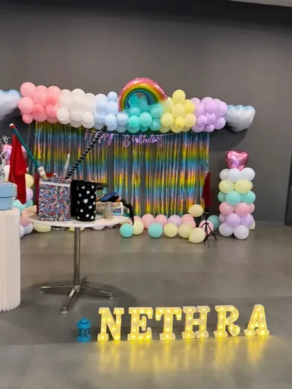 A wide shot of a rainbow-themed party setup, complete with a balloon garland, shimmer backdrop, and marquee letters.