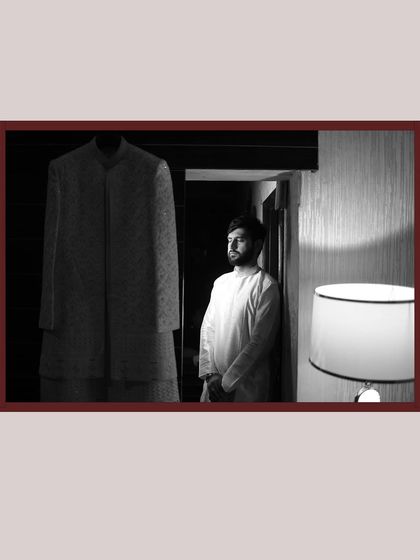 An artistic black and white shot of the groom in a moment of quiet contemplation before getting dressed, with his sherwani hanging nearby.