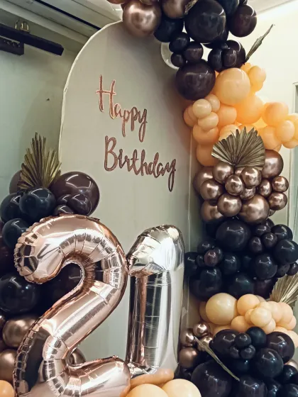 A trendy and modern decoration for a 21st birthday. This setup uses an arched backdrop, large foil number balloons, and a sophisticated balloon arrangement in black, brown, and peach tones.