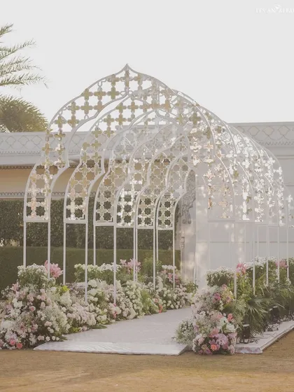 The grand entrance tunnel for a Mughal-themed wedding. The intricate white jali arches, lined with pastel flowers, create a breathtaking walkway for guests arriving at the ceremony.