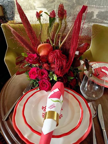 A place setting featuring a custom-printed napkin and a small, sculptural arrangement of red roses and dried botanicals. This shows how we carry the central theme through to each guest's individual space.
