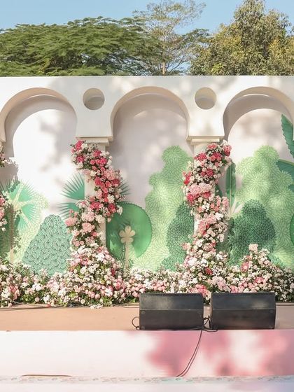 A beautiful photo backdrop featuring white arches and a hand-painted mural of a lush, stylized forest, with pillars wrapped in spiraling pink and white floral garlands.