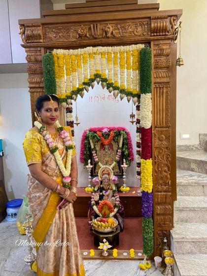 A client celebrating her housewarming ceremony. She is dressed in a traditional silk saree with elegant makeup, standing before her decorated home temple.
