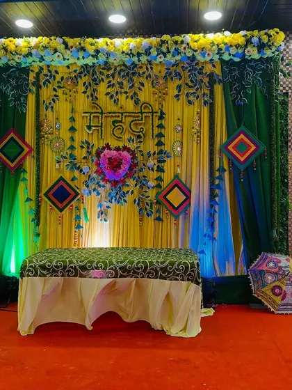 A stunning Mehendi stage with a rich green and yellow backdrop, decorated with floral cutouts, a heart-shaped flower arrangement, and colorful hangings.