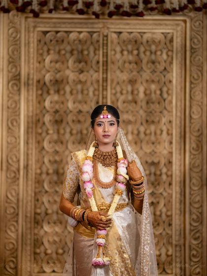 A portrait of the bride against a beautifully carved wooden door. Her white and gold saree, paired with a lotus garland, creates a look of traditional purity and grace.