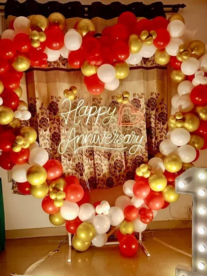 Celebrating the first year of togetherness with a beautiful at-home setup. This heart-shaped balloon arch in red, white, and gold is paired with a '1' marquee number and a "Happy Anniversary" neon sign.