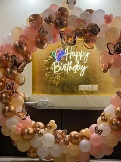 A soft and pretty ring backdrop for a home celebration. The arch of rose gold, peach, and white balloons is decorated with butterfly accents and a multi-color neon sign.