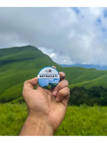A hand holding the Netravati trek badge against the backdrop of the iconic peak.