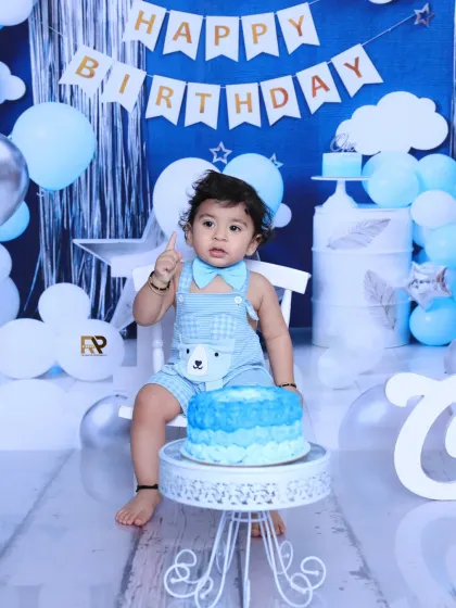 Another adorable shot from the blue-themed cake smash, with the birthday boy pointing up, full of excitement. The setup includes a birthday banner, balloons, and a stylish cake stand.
