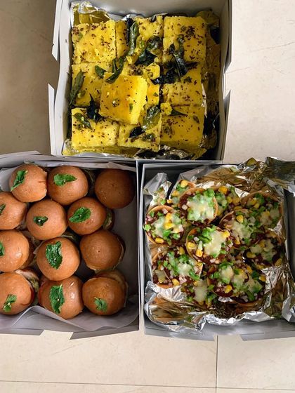 Boxes of freshly prepared Khaman Dhokla, mini veggie burgers, and pizza crackers ready for delivery. A perfect combination of Indian and continental snacks.