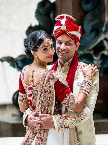 An elegant couple posing together. The groom is in a classic cream sherwani with red and gold trim, complemented by a red and white safa.