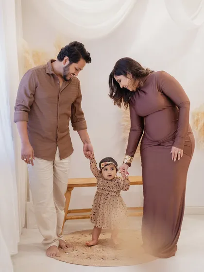 A family of three, celebrating their daughter's first steps and first birthday. The coordinated brown outfits and minimalist boho set create a warm, earthy-toned family portrait.