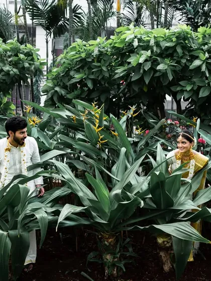 A playful moment during a South Indian wedding portrait session, with the couple peeking at each other through the plants.