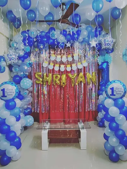 A "First Birthday Boy" themed decoration in shades of blue and white. This full room setup includes a balloon arch, shimmer backdrop, and multiple themed foil balloons to celebrate the special milestone.
