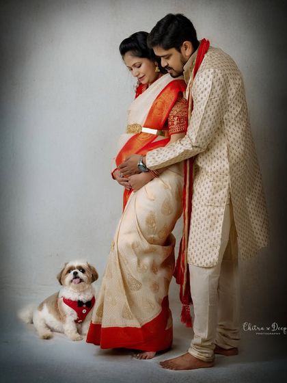 A classic maternity portrait featuring an expecting couple in traditional Indian wear, with their adorable dog sitting at their feet.