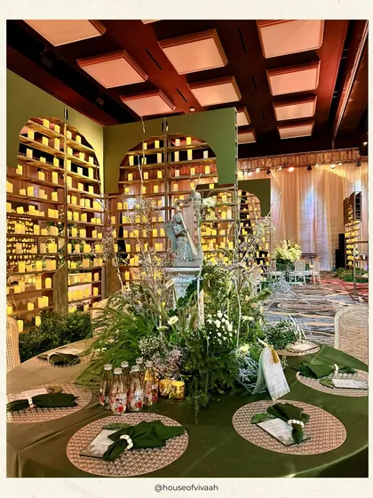 A dining table at a greenhouse-themed wedding, featuring an olive green tablecloth, woven placemats, and a centerpiece with a classical bust, all set against a wall of candles.