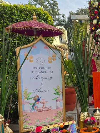A personalized welcome sign for Avani and Aanjney's Sangeet. The design features beautiful illustrations of peacocks and fountains, topped with a traditional Indian umbrella for a royal touch.