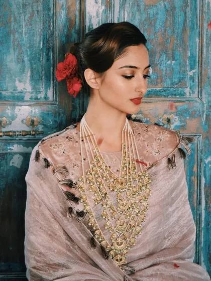 A full view of the vintage-inspired festive look. The model wears a beautiful Payal Singhal outfit with a layered Tyaani jewellery necklace, with red roses in her hair adding a touch of classic romance against the rustic blue door.