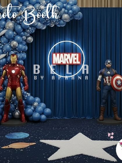A dedicated photo booth for the Marvel theme, featuring life-size statues of Iron Man and Captain America. The neon Marvel logo and blue drapery provide a clean, powerful backdrop for superhero poses.