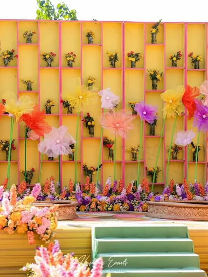 A creative and colorful haldi stage backdrop. The yellow wall features a grid of small floral arrangements, with large, whimsical paper flowers adding a touch of fantasy.