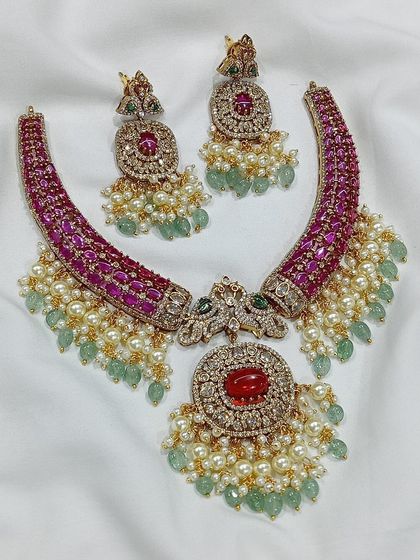 This antique-finish necklace set features peacock motifs and intricate ruby and green stone work. It comes with matching earrings and is perfect for customers who appreciate detailed, traditional designs.
