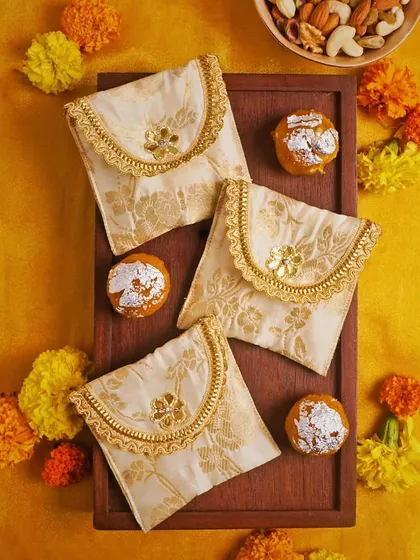 A festive flat lay featuring elegant cream and gold pouches on a bright yellow background with marigold flowers and ladoos. This image is rich with cultural context, perfect for Diwali or other Indian celebrations.