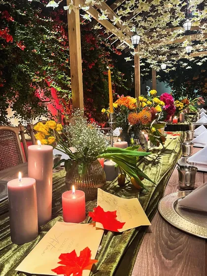A long dining table set for an autumn-themed dinner. The decor features a deep green velvet runner, candles in warm tones, and colorful fall foliage, creating a cozy and inviting ambiance.