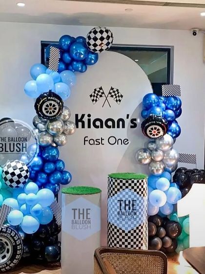 A "Fast One" race car theme for a first birthday. The decor features a custom checkered flag backdrop, balloon arches in shades of blue, and car-themed props.