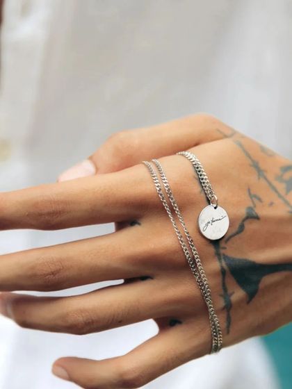 A man holding a silver-toned round pendant with a custom script engraving. The shot against his tattooed hand creates a modern and edgy look.
