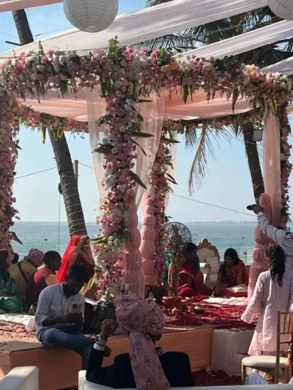 A candid moment from a beautiful daytime beach wedding. The mandap is wrapped in pastel pink flowers and sheer drapes, creating a light and airy feel that complements the sunny seaside location.