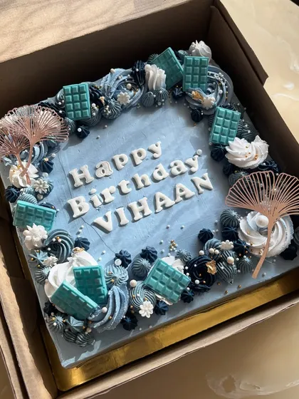 A sophisticated square birthday cake in shades of blue and white. It's decorated with piped buttercream, mini chocolate bars, and elegant fan toppers for a modern look.