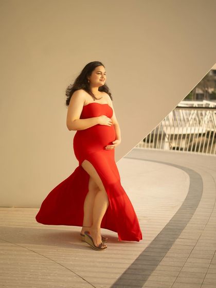 A beautiful profile shot that highlights the baby bump against a clean, architectural background. The slit in the red dress adds a touch of modern style to this outdoor portrait.