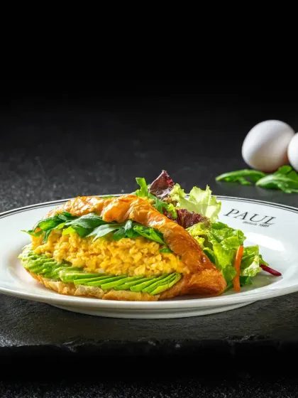 A gourmet scrambled egg and avocado croissant sandwich, elegantly plated and shot against a dark, moody background for Paul.