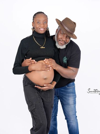 A sweet and natural couple portrait with the husband-to-be showing his affection. Their casual black outfits and the simple white background create a relaxed and authentic feel.