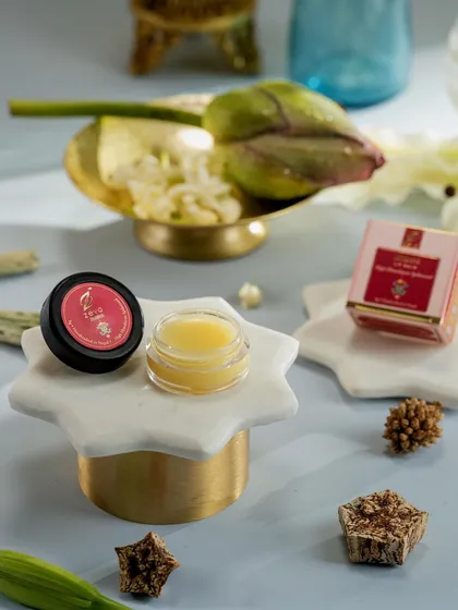 A clean and artistic shot of Zeva's lip balm, showing the product open to reveal its texture and color. The use of marble, brass, and fresh botanicals adds a touch of modern elegance.