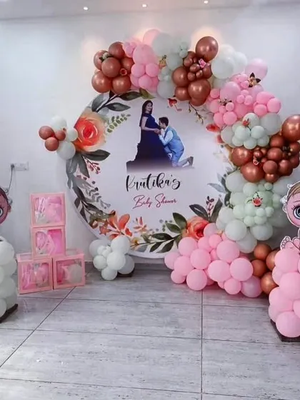 A personalized baby shower decoration featuring a large circular backdrop with the parents-to-be's photo. The setup is framed with a beautiful balloon arch in dusty rose, pink, and white, with cute baby character cutouts.