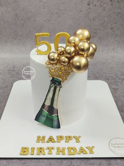 A minimalist champagne bottle cake. This clean, white design features a champagne bottle popping with golden spheres, perfect for a 50th birthday.