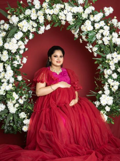 A festive and joyful studio setup with the mom-to-be in a stunning red gown, seated within a beautiful arch of white flowers.