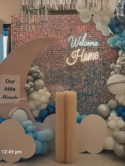 A space-themed welcome home decoration with a silver sequin backdrop. The design features a moon cutout, planet-like balloons in shades of blue and silver, and a 'Welcome Home' neon sign for a futuristic and cool look.