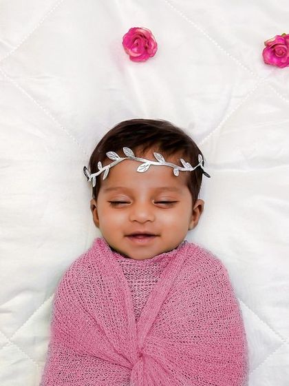 A happy, smiling baby wrapped in pink and wearing a leafy crown, surrounded by scattered rose petals on a white quilted background.