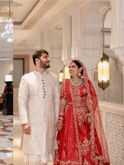 A classic wedding portrait in a grand hotel corridor. The couple's traditional red and white attire looks stunning against the architecture.