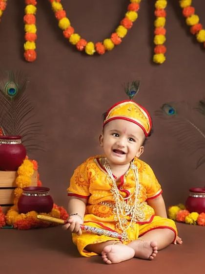 A smiling baby dressed as little Krishna sits in a festive Janmashtami setup with marigolds and peacock feathers.