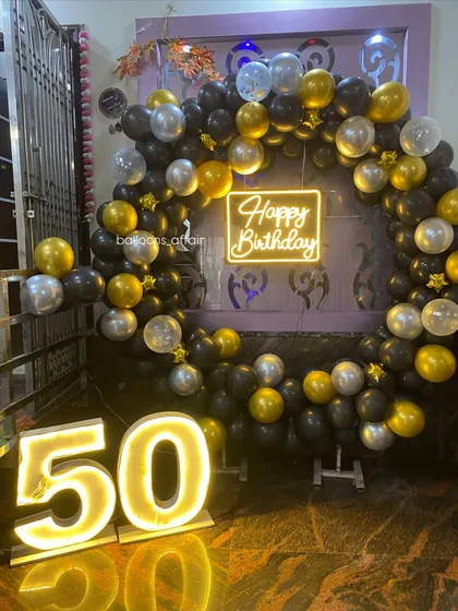 Celebrating a 50th birthday in style with a glamorous black, gold, and silver theme. This setup includes a full circular balloon arch, a neon 'Happy Birthday' sign, and large illuminated marquee numbers for a milestone event.
