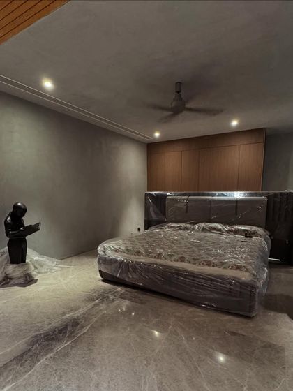 A work-in-progress shot of a bedroom with a moody aesthetic. The space features micro-concrete walls, a custom upholstered bed, and built-in wooden wardrobes, all awaiting final touches.