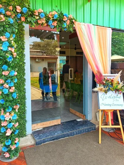 A welcoming entrance for a naming ceremony. The doorway is decorated with a floral arch in blue and peach, with a sign on an easel that says 'Welcome to the Naming Ceremony'.