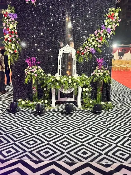 A chic outdoor stage setup with a black shimmer backdrop, elegant floral arrangements, and a unique geometric patterned floor.