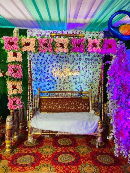A close-up of a baby shower swing (jhula) decorated with pink flowers and set against a green backdrop with a neon sign.