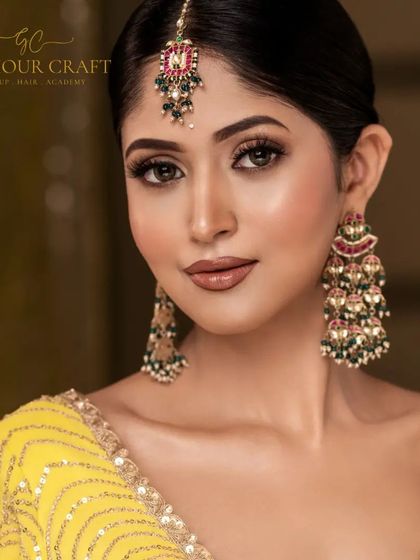 A close-up portrait for a Haldi or Mehendi look. The focus is on the glowing skin, the statement earrings, and the maang tikka.